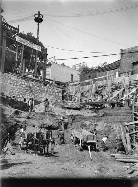 Construction site showing men excavating the foundation and basement of a building. Teams of horses are hitched to wagons. The men are using various implements. Wood scaffolding is holding up the sides of the excavation and a stone foundation is exposed at the back. Other buildings are in the background. A banner above the stone foundation reads: "Shobis Bros. Structural Steel Works, Milwaukee — Wisc."