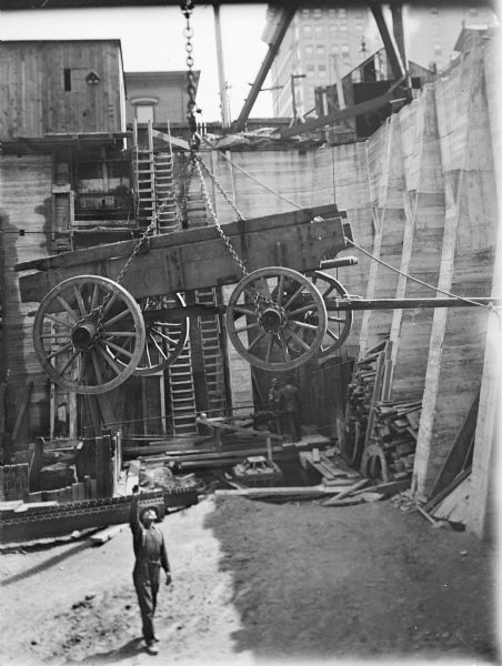 A wagon is being raised or lowered at a construction site. The wagon has chains looped around the axle of each wheel as a man below signals directions. He is standing in the bottom of a large excavated foundation with concrete walls. Two other workers are standing near the far wall with a number of long ladders.