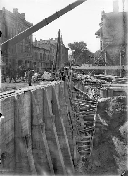 A construction site with a view of the excavated foundation and the street level. At the street level are buildings, people, and horse-drawn wagons. One man stands close to the edge, holding a rope tied to a chain, attached to a steel beam. The foundation has concrete supports on the street side. Scaffolding, lumber and barrels can be seen on the right.