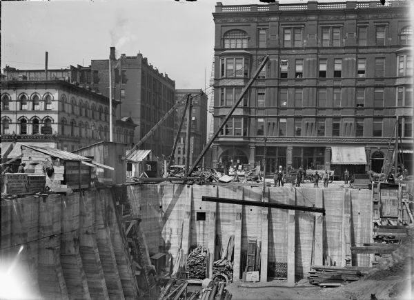 A construction site with a view of the excavated foundation and the street level. At the street level are buildings, people, a crane, construction shacks, material and horse-drawn wagons. A crane lowers or raises a steel beam. The foundation has concrete supports. Scaffolding, lumber and ladders can be seen in the excavated foundation.