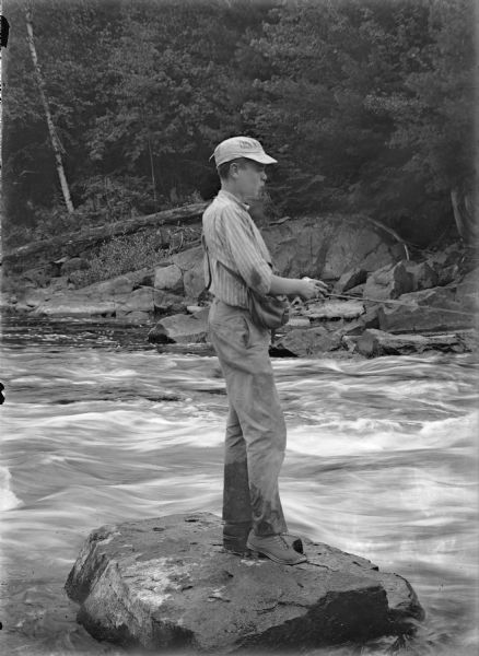 A man is fishing while standing on a large, flat rock in a river. In the background along the shoreline are rocks and trees.