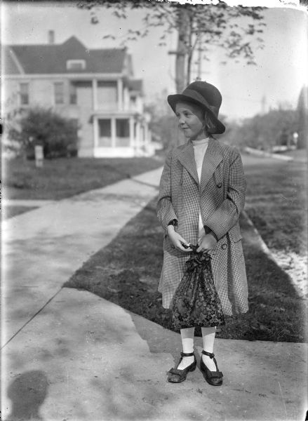 A girl is standing on a sidewalk, gazing to the left. She is wearing a coat, hat, stockings and shoes with bows. She is carrying a flowered cloth drawstring bag. In the background is a residential street with houses.