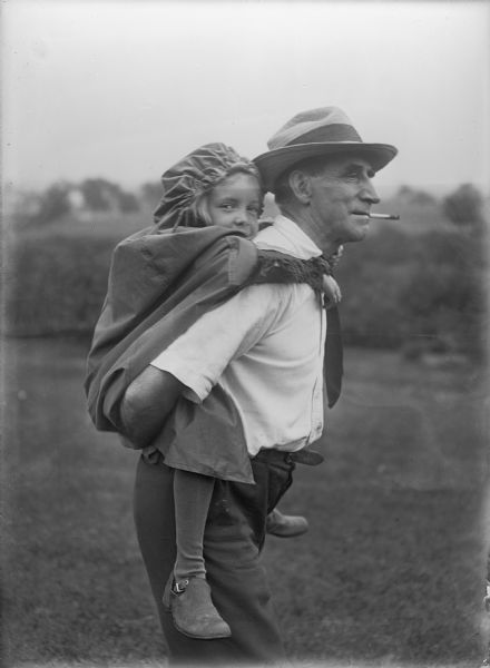 A man is carrying a girl piggyback outdoors while smoking a cigarette. They are standing on a lawn with trees in the background. She is wearing a dress, coat, cap, stockings and shoes. He is wearing a short-sleeved shirt, trousers, belt, necktie and a straw hat with a band.