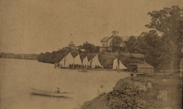 View along the Lake Monona shoreline towards the Tonyawatha Spring Hotel. A crowd of people are standing on the pier with several sailboats. On the shore is a boathouse. In the lower left is someone paddling a canoe. The hotel, on the east shore of the lake, was located in Blooming Grove Township, just outside of Madison. The hotel opened in 1879. On July 31, 1895, the hotel was destroyed by a fire.