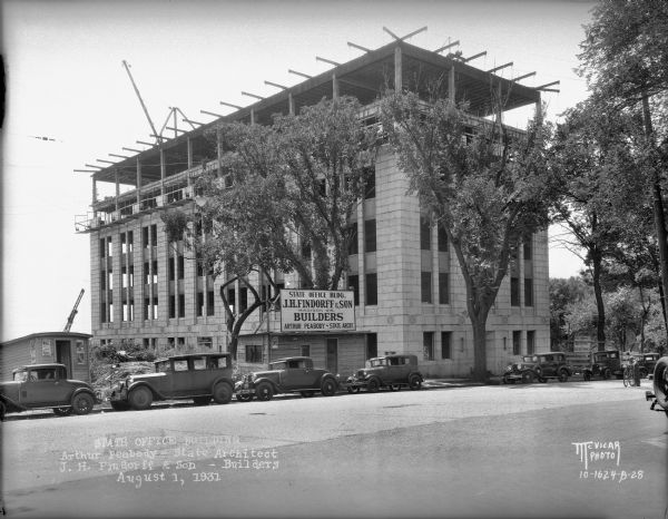 State Office Building, 1 W. Wilson Street, under construction. View from the left on Wilson Street with automobiles and a bicycle in the foreground. The sign reads: "State Office Bldg., J.H. Findorff & Son Builders, Madison Wis., Arthur Peabody-State Archt."