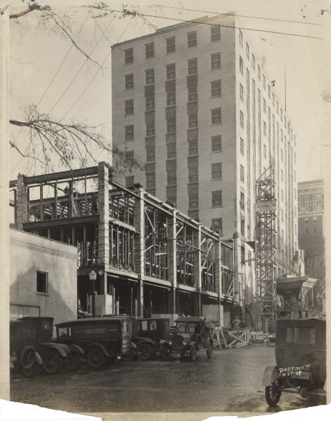 Construction site of the Wisconsin Power and Light Co. Office Building & Bus Station. The address of the bus station was 122 West Washington Avenue, but the entrance was on North Fairchild Street. A sign on the scaffold reads, Wisconsin Power and Light Co. Office Building & Bus Station, J.H. Findorff & Son Builders, Law, Law & Potter, Archts." The tall building was the Wisconsin Power & Light Building, bought in 1970 by the Hovde Properties. On the left is Arcade Tailors and Cleaners with their trucks parked in front. In the background is the Hotel Loraine, currently condominiums. 