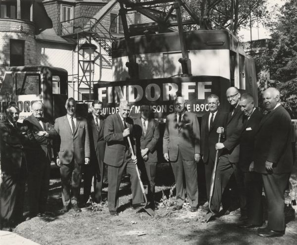 Eleven men are standing during a groundbreaking ceremony in front of a Findorff crane. Two men are holding shovels. The sign painted on the crane reads: "Findorff, Builders of Madison Since 1890." 