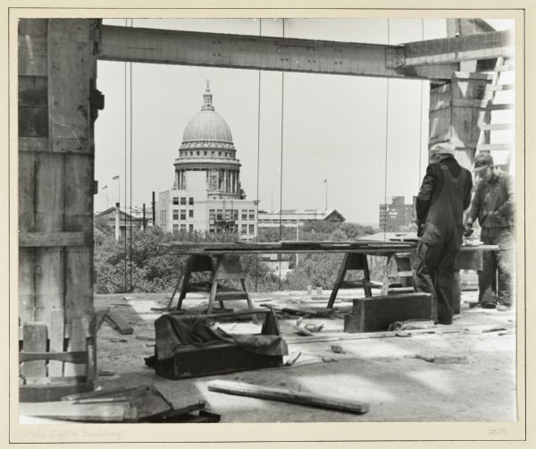 A State Office Building construction site, downtown, with a view of the Wisconsin State Capitol. Two men on the right are working with boards on sawhorses. On the floor are tools, toolboxes and more wood, and above are steel girders.