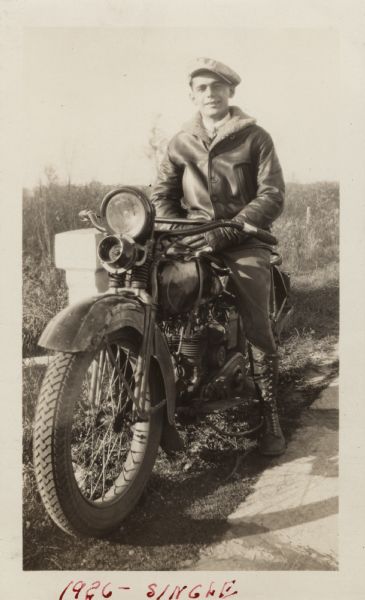 Raymond (Ray) Griesemer is sitting on his Harley-Davidson motorcycle. He is wearing leather gloves, a jacket with sheepskin lining, dark pants, lace-up boots and a cap. The motorcycle is in front of a stone wall. In the background is a field with grass, weeds and trees. Handwritten at the bottom: "1926 — Single."