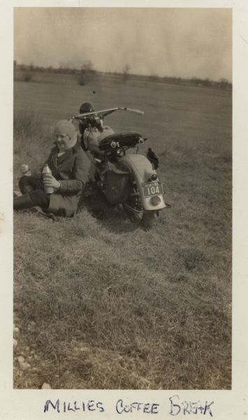 Mildred (Millie) Griesemer is sitting in the grass on the ground against the side of her Harley-Davidson motorcycle, holding a thermos and cup. She is wearing a dark jacket, pants, and boots. Her sweater and hood are light-colored. The motorcycle is parked in a field, and in the far background are trees. The license plate reads: "WIS-35, 104." Handwritten at the bottom: "Millies coffee break."