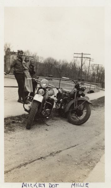 Two Harley-Davidson motorcycles are parked next to a sidewalk on the side of a dirt road. Just behind the motorcycles are a man and two women standing together while leaning against a metal railing. The woman on the right is Mildred (Millie) Griesemer. They are all wearing jackets, pants and lace-up boots. The man is wearing a leather helmet and goggles, and the women are wearing tight fitting caps. Below the railing is water, perhaps a river, and in the background an automobile is moving across a bridge. On the opposite shoreline are trees and a large building. Handwritten at the bottom: "Mickey, Dot, Millie."