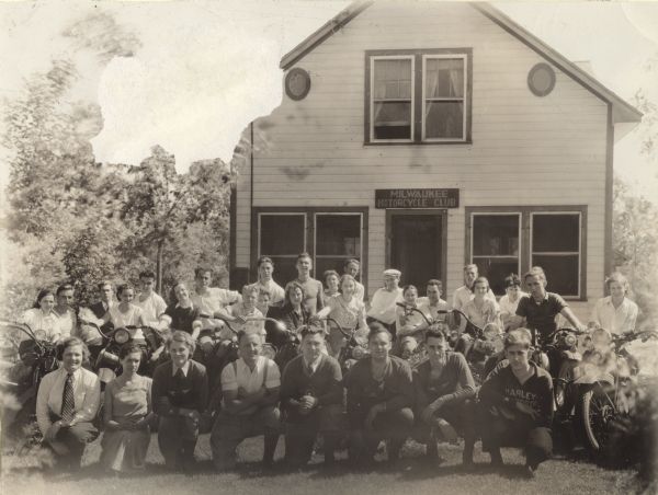 Outdoor group portrait of men and women posing on the lawn in front of their clubhouse. The front row of the group is kneeling, the middle row is sitting on motorcycles, and the back row is standing. 6th and 7th from the left are Mildred (Millie) and Raymond (Ray) Griesemer. Several people are wearing Harley sweaters. Above the door is a sign that reads: "Milwaukee Motorcycle Club."