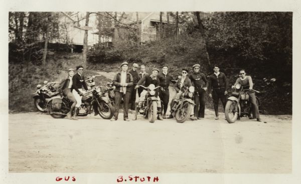 Informal group portrait of eleven motorcycle riders. Some of the motorcycles are Harley-Davidsons. Most of the riders are standing, wjith some are sitting on their motorcycles. They are all dressed in riding clothes. A gravel area is in the foreground, and in the background is a steep bank covered with trees and foliage, with houses at the top. Handwritten at the bottom: "Gus" and "B. Stuth."