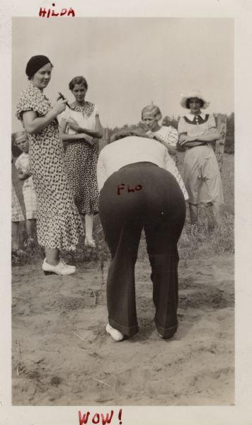 A group of women and girls wearing dresses are standing and gazing at Flo Knuth, who is wearing trousers. Flo is standing and bending over in the foreground, showing her posterior towards the camera. "Flo" has been handwritten in red ink on her behind. Handwritten at the top: "Hilda," and handwritten at the bottom: "WOW!"