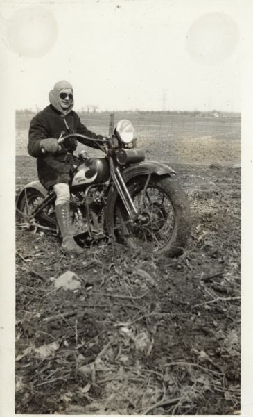 A man is sitting on his Harley-Davidson motorcycle in a muddy field. The back wheel is partially buried in the mud. He is wearing leather gloves and a coat with a lining, dark pants, lace-up boots, a hood and sunglasses. In the background is a fence and a field.