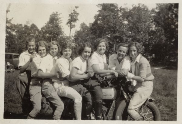 Eight women are perching on and around a Harley-Davidson motorcycle. They are all wearing light-colored blouses and jodhpur style pants with boots. In the background is a grassy field, trees and automobiles.