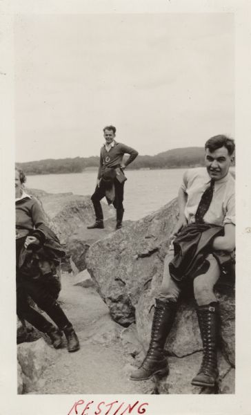Three motorcycle riders are posing on boulders along the shore of Devil's Lake. One rider (a woman?) is partially out of the frame on the left. Elmer, right, is wearing a shirt, tie, trousers and high lace-up boots, and is holding a leather jacket. The man in the center in the background is Raymond (Ray) Griesemer, who is wearing a sweater with the Milwaukee Motorcycle Club patch on it. In the background is the lake and far shoreline. Handwritten at the bottom: "Resting."