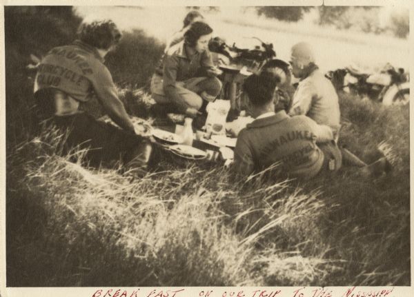 Milwaukee Motorcycle Club members at an outdoor breakfast in a field next to the road. Several members are wearing jackets with "Milwaukee Motorcycle Club" on the back. The closest man with his back to the camera may be Raymond (Ray) Griesemer. Food and dishware are arranged in the middle. Harley-Davidson motorcycles are parked on the edge of the road. Handwritten at the bottom: "Breakfast on our trip to the Mississippi."