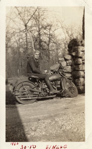 A man is sitting on his Harley-Davidson motorcycle parked along a gravel road near a stone wall. He is wearing leather gloves, a jacket, dark pants, lace-up boots and a leather cap with goggles. In the background is a field with grass and trees. Handwritten at the bottom: "1930 30-50 Single."