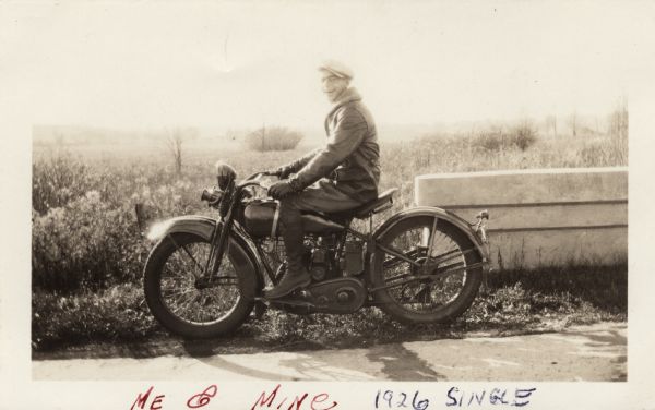 Raymond (Ray) Griesemer is sitting on his Harley-Davidson motorcycle. He is wearing leather gloves, a jacket with a sheepskin lining, dark pants, lace-up boots and a cap. The motorcycle is in front of a stone wall. In the background is a field with grass, weeds and trees. Handwritten at the bottom: "Me & Mine, 1926 Single."