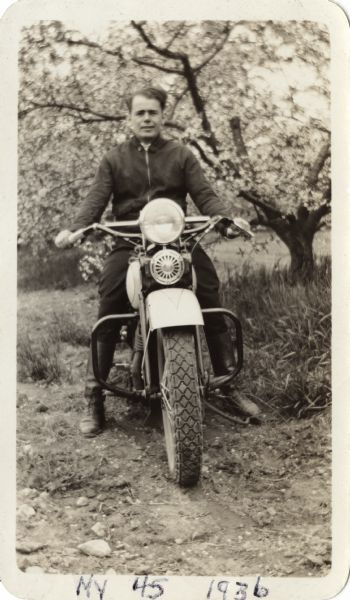 Raymond (Ray) Griesemer is sitting on his Harley-Davidson motorcycle. He is wearing a jacket, dark pants and boots. In the background is a field with grass and trees. Handwritten at the bottom: "My 45 1936."