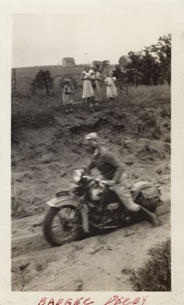 A man is riding his Harley-Davidson motorcycle in the Badger Derby. He is racing through a ditch filled with sand, wearing motorcycle gear. On the opposite bank, a group of women and girls, wearing dresses, are watching. In the background are fields, trees and farm buildings. Handwritten at the bottom: "Badger Derby."