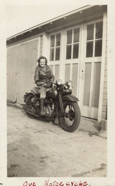 Mildred (Millie) Griesemer is sitting on her Harley-Davidson motorcycle in front of a garage door, parked in an alley. She is wearing leather gloves and jacket, dark pants and lace-up boots. Handwritten at the bottom: "Our Motorcycle."