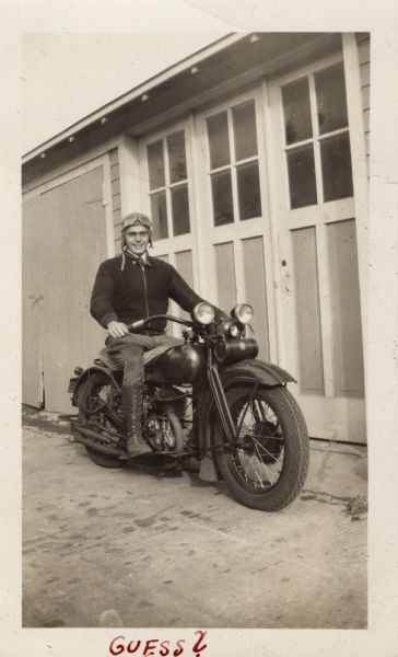 Raymond (Ray) Griesemer is sitting on his Harley-Davidson motorcycle in front of a garage door. He is wearing a  jacket, pants, lace-up boots and a leather cap with goggles. Handwritten at the bottom: "Guess?"