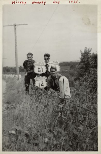 Three men are standing and posing with Minnesota highway signs near a post and cable guard rail. US 61 and US 14 are on two signs. They men are wearing motorcycle gear, and are surrounded by grass, weeds and shrubs. Raymond (Ray) Griesemer is the man standing on the left, Harvey is standing on the right, and Mickey is kneeling in front. A power pole is in the background. Handwritten at the top: "Mickey, Harvey, Ray, 1935."