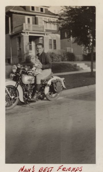 View across street towards Raymond (Ray) Griesemer is holding a dog in his lap while sitting on a Harley-Davidson motorcycle parked at the curb. He is wearing jacket, pants and boots. There are houses in the background. Handwritten at the bottom: "Man's Best Friends."