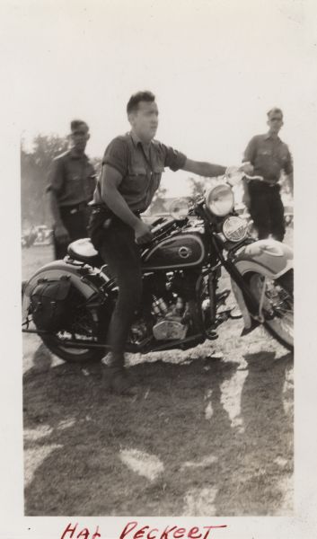 Harold (Hal) Deckert is straddling his Harley-Davidson motorcycle, which is on a grassy hill. He is wearing a shirt, dark pants and boots. In the background two men are standing and watching him. Hal was a test rider for Harley-Davidson. Handwritten at the bottom: "Hal Deckert."