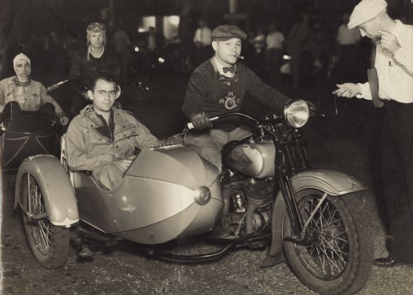A man, on the right, is riding a Harley-Davidson motorcycle with a man in a sidecar, on the left, in an event for the Badger Derby. The driver who is smoking a cigar is wearing a sweater with a Milwaukee Motorcycle Club logo on it, bow tie, driver's cap. The passenger is Raymond (Ray) Griesemer, who is wearing a jacket and pants. Two more men riding a motorcycle with a sidecar are behind them. On the right, a man is standing and looking at a stopwatch. In the background are spectators. A family story indicates that the original passenger, a woman, fell ill and Ray was the next lightest person in the crew. He became the passenger.