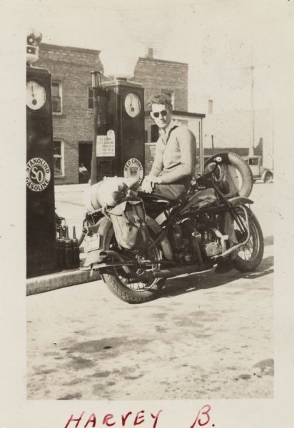 Harvey B. is sitting sideways facing backwards on his Harley-Davidson motorcycle at a service station. He is wearing a sweater with the Milwaukee motorcycle Club patch, dark pants and sunglasses. In the background are two gas pumps, Stanoline and Red Crown, a man sitting in the open doorway of the service station, and an automobile. Handwritten at the bottom: "Harvey B."