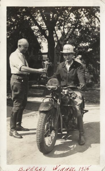 The 1935 Badger Derby winner, John T. Chasty, receiving his trophy. He is sitting on his motorcycle and is wearing motorcycle gear and a special derby hat. The presenter is standing on the left, who is also wearing motorcycle gear and a necktie. In the background are trees and buildings. Handwritten at the bottom: "B. Derby Winner 1935."