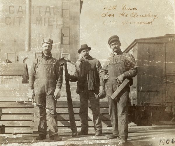 Three men are posing in front of construction materials. The man on the left is holding a hand drill, the man in the center is holding a long saw, and the man on the right is holding a carpenters' plane. There is a building in the background with a sign that reads: "Capital City Mill." There is a railroad car on the far right behind the men. Caption reads: "Bill Dun, Chas. McClousky, and Bill Hamond."