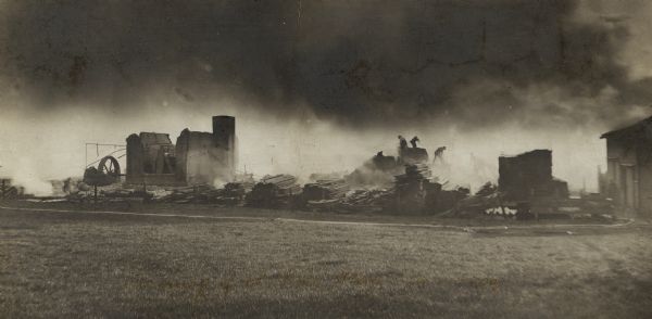 View across field towards buildings smoking after the fire. Men are standing on and around stacks of lumber. Caption reads: "The Morning After the Fire."