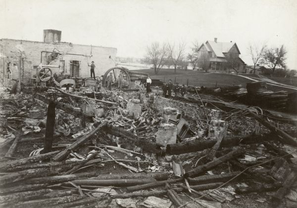 Elevated view of the damage from the fire. Burned logs are in the foreground, and pieces of burned and broken machinery is among the broken foundation. Men are standing near the brick building behind the wreckage, and nearby are stacks of lumber. In the far background is a lawn and a large building with a porch near the shoreline of a lake. Caption reads: "On Friday, the 13th 1909, the Findorff Plaining [sic] Mill and Lumber Yard were completely destroyed by fire resulting in a loss in excess of $100,000; insurance coverage was about $17,000."