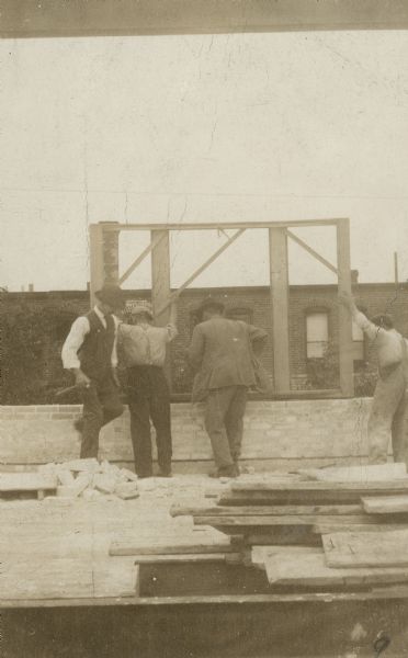 Three men are working on the top of a building, putting a wooden structure on top of a brick wall. Stacks of lumber are in the foreground. The top of a brick building is in the background. Caption reads: "J.H. Findorff (with hammer) working during a strike."