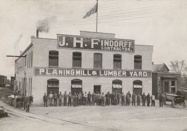 Elevated group portrait of men posing in front of the J.H. Findorff, Contractor, Planing Mill & Lumber Yard building. A smaller brick building on the far right has a sign that reads: "Office." Railroad tracks are in the foreground, and railroad cars are on the tracks in the background on the left. Caption reads: "Just a year after the fire J.H. Findorff completed the new building that was to house his business on the same location on W. Wilson St."