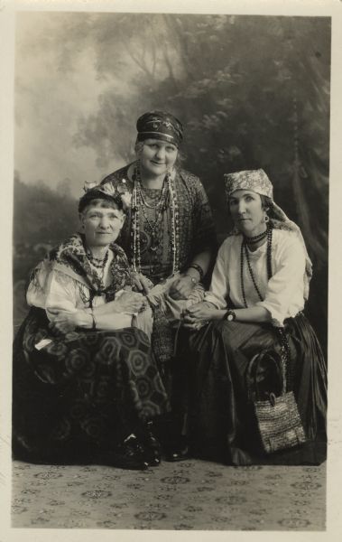 Full-length studio portrait in front of a painted backdrop of three women wearing gypsy costumes. One women is sitting in the center, and two women are sitting next to her on the left and right. The women are dressed in skirts, blouses, jewelry, shawls and headscarves or hats. Two of the women have handbags. The Gypsy Party was an Eastern Star event. Names (l to r) Jennie White, Emma W. Gebhardt and Geda (Gyda) Severson. There is a patterned carpet beneath them. <p>Part of a series of portraits taken of guests at an Eastern Star "Gypsy Party."</p>