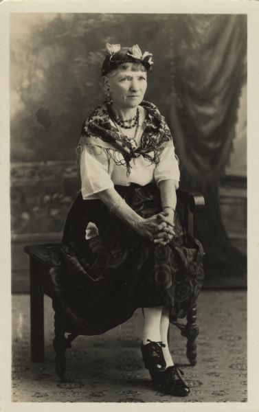 Full-length studio portrait of Jennie White sitting on a chair in front of a painted backdrop. She is wearing a long, dark, patterned skirt, a light blouse, shawl, flowers in her hair, earrings, and many necklaces and bracelets. Her hands are clasped on her knees.<p>Part of a series of portraits taken of guests at an Eastern Star "Gypsy Party."
