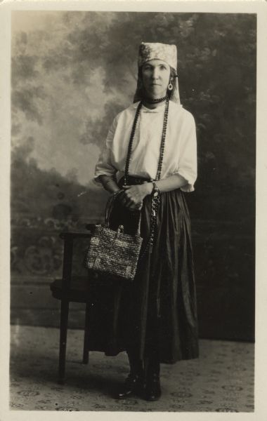 Full-length studio portrait of Geda (Gyda) Severson standing next to a chair in front of a painted backdrop. She is wearing a long, dark skirt, a light blouse, headscarf, earrings, necklaces and bracelets. She is holding a handbag in both hands. <p>Part of a series of portraits taken of guests at an Eastern Star "Gypsy Party."