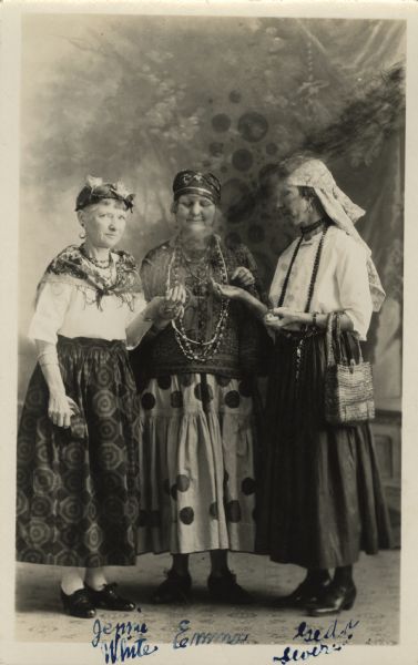 Full-length studio portrait in front of a painted backdrop of three women wearing gypsy costumes. The woman in the center is gazing at the other women's palms that they are holding up to her. The women are dressed in skirts, blouses, jewelry, shawls and headscarves or hats. The woman on the right has a handbag over her arm. The Gypsy Party was an Eastern Star event. Names (l to r) Jennie White, Emma W. Gebhardt and Geda (Gyda) Severson. They are standing on a patterned carpet. <p>Part of a series of portraits taken of guests at an Eastern Star "Gypsy Party."</p>