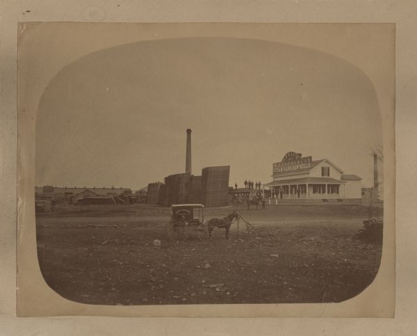 Albumen print of the office of the W.J. Young & Co. Steam Gang Saw Mill. Several people are standing on the porch and a nearby wooden structure. A horse-drawn wagon stands in front of the office. Another horse-drawn wagon in the foreground has a sign on the side that reads: "Northwestern View Co., 5." In the center is a smokestack behind large piles of sawn boards. On the left are more buildings and wood piles.