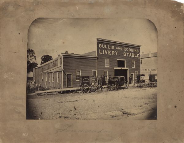 Albumen print of a livery stable on an unpaved street with wooden boardwalks. The name of the stable is "Bullis and Robbins. Livery Stable." A sign on the building to the right reads "Livery Stable." Five men are standing and two are lounging on the boardwalk left of the door. One man stands to the right. Parked in front is an enclosed wagon with "Northwestern View Co. No. 5" painted on the side and back. Three buggies are parked to the right. The vehicles are horse-drawn but the horses are not hitched up. Below the image is hand written "Fond du Lac."