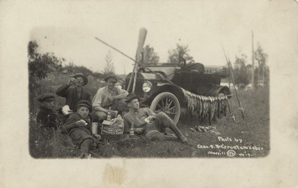 Real Photo Post Card of a group of fishermen taking a food and beverage break. Three men and two youth are lounging on the ground with bottles and sandwiches. A "Queen Assortment Fancy Cookies" box in the middle of the group probably contains the food and drink. They are wearing casual clothes and hats. The car is draped with fish strung on a pole, fishing poles and oars. More fish on a stringer lay on the grass. In the background are a lake, hill, foliage and trees. "Photo by Geo. O.B. Gruetzmacher, LTD, Merrill Wis." is written on the negative.