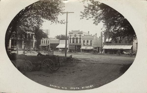 Real Photo Post Card of a South Main Street scene. Men and women walk along the sidewalks and many horse-drawn vehicles are tethered in front of the stores. The streets are not paved, on the left is an area with horses, wagons and buggies. One wagon has "New Stoughton Wagon" painted on it. A sign "Howe" and "Borden & Selleck[?]" is set between the street and parking area. Retail and commercial buildings line the street. Signs on the buildings read, "Oregon Drug Co, Drugs, Jewelry, Silverware & Crockery," "A.M. Anderson Groceries," "B. Mc Dermott Sons" and "1898" on the Masonic Lodge. On the left, behind the trees, are many businesses inside the Netherwood Building. Caption Reads: "Oregon, Wis."