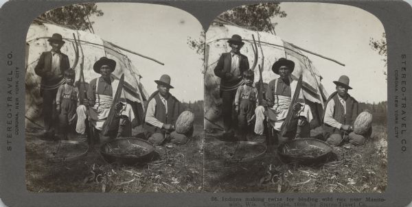 Stereograph of three Native American men and a child, outdoors, in front of a wigwam. On the ground is a basket of materials. The man on the left is standing and holding a knife and two pieces of bent wood. Next to him is the child, holding a wooden tool. The center man is holding a rifle and possibly a stretched animal skin on a board. The man on the right is winding the twine into a large ball. Caption on bottom right reads: "56. Indians making twine for binding wild rice near Manitowish, Wis."