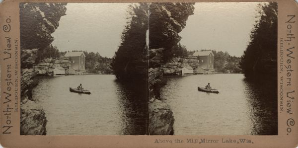Stereograph of a man fishing from a rowboat in the millpond above Timme's Mill on Mirror Lake. The boat is framed by rock formations and tree on both sides. In the background are the flour mill, two boathouses and the dock. Caption at foot: "Above the Mill, Mirror Lake, Wis."