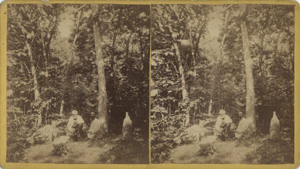 Stereograph of three men having a picnic in a clearing in the woods at Devil's Lake or nearby. One man is sitting against a tree, another is kneeling and the last man is reclining. Containers, baskets and bottles are holding their food and drink. Mature trees are towering over the site. 